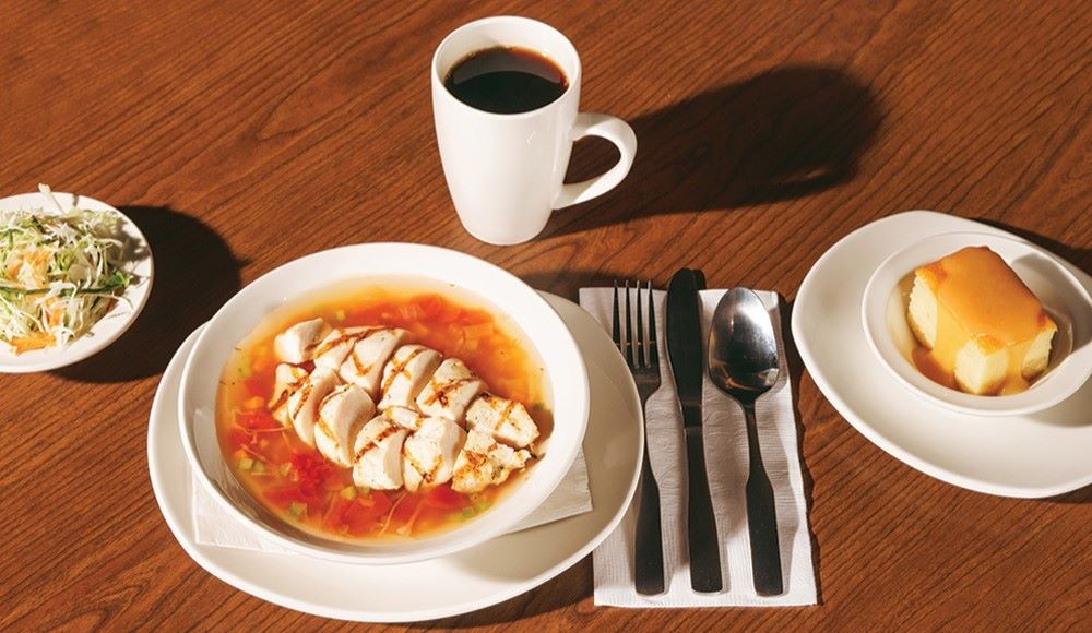 Sur une table, un repas complet du midi St-Hubert avec la soupe-repas au poulet et aux légumes, un café, une salade de choux traditionnelle et un gâteau vanillé.